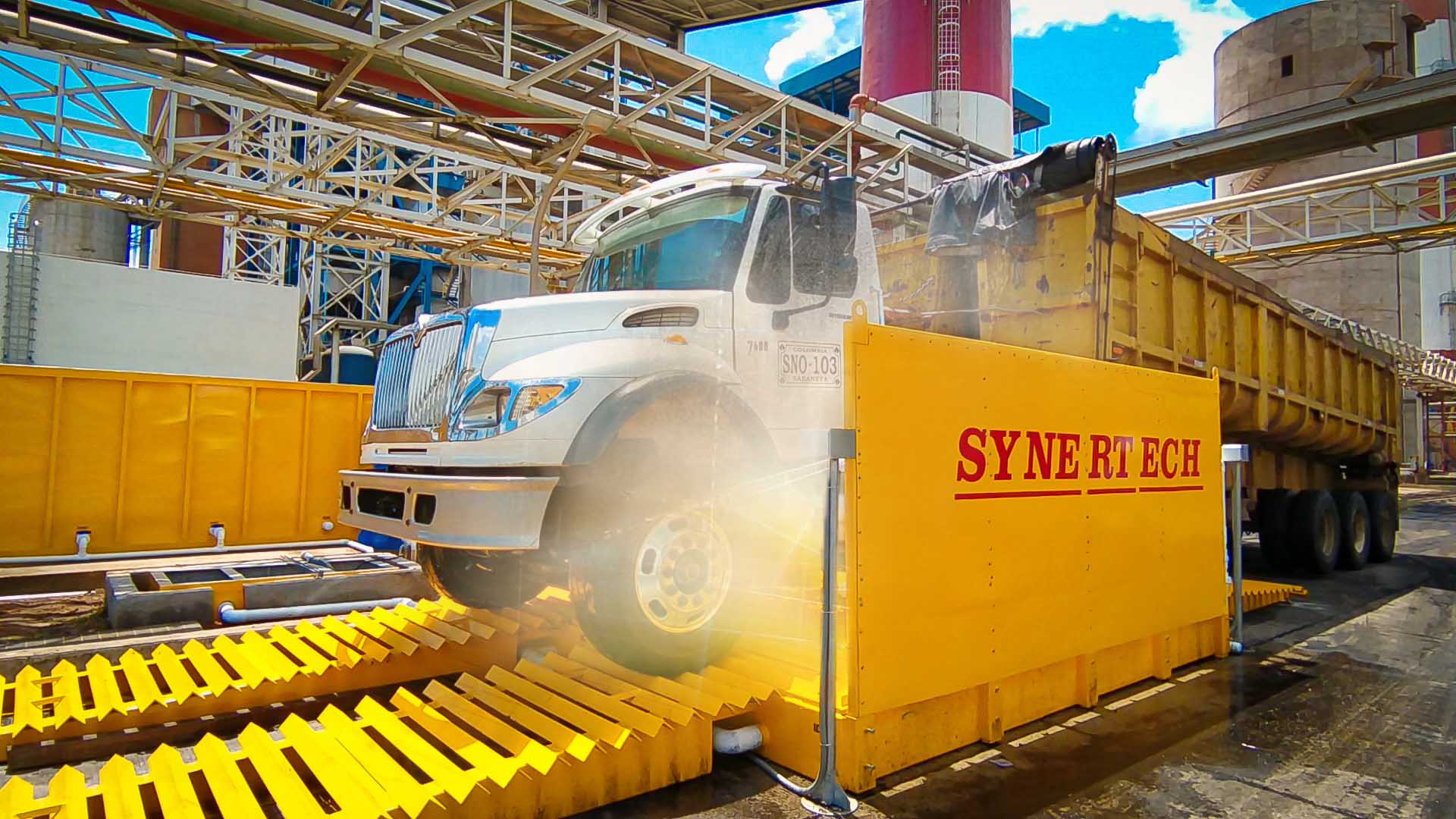 Camión grande blanco y amarillo pasando por un sistema de lavado de ruedas activo, rociando agua para limpiar la suciedad y escombros. En un sitio industrial o de construcción con silos al fondo, el sistema ayuda a mantener limpias las vías públicas. - ver más