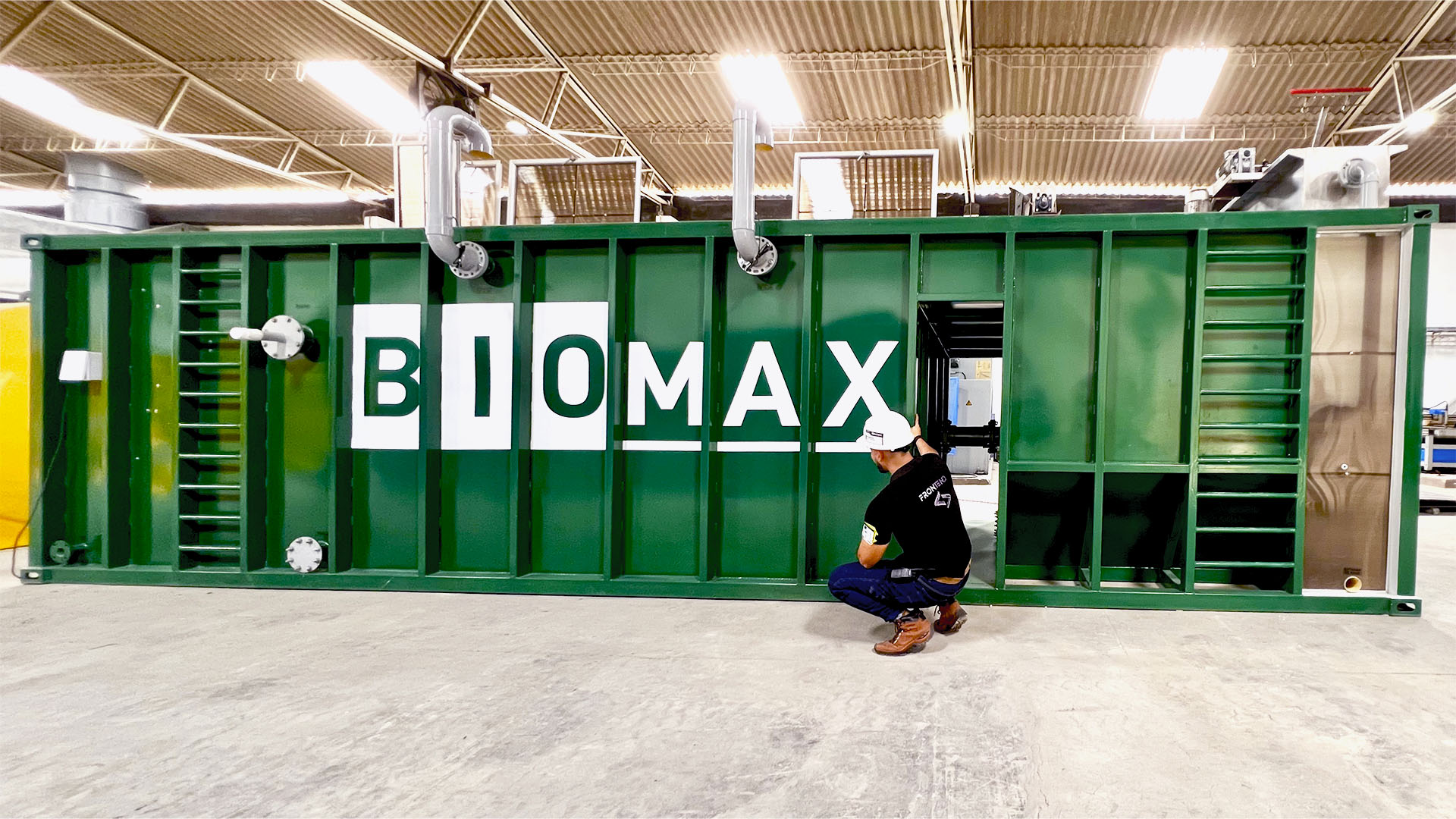 Técnico inspeccionando una planta de tratamiento de aguas residuales modular de color verde marca BIOMAX SYNERTECH, que incluye módulos de pretratamiento, decantación, DAF y oxidación avanzada. La planta está ubicada en un entorno industrial con techo alto y tuberías visibles. - ver más información