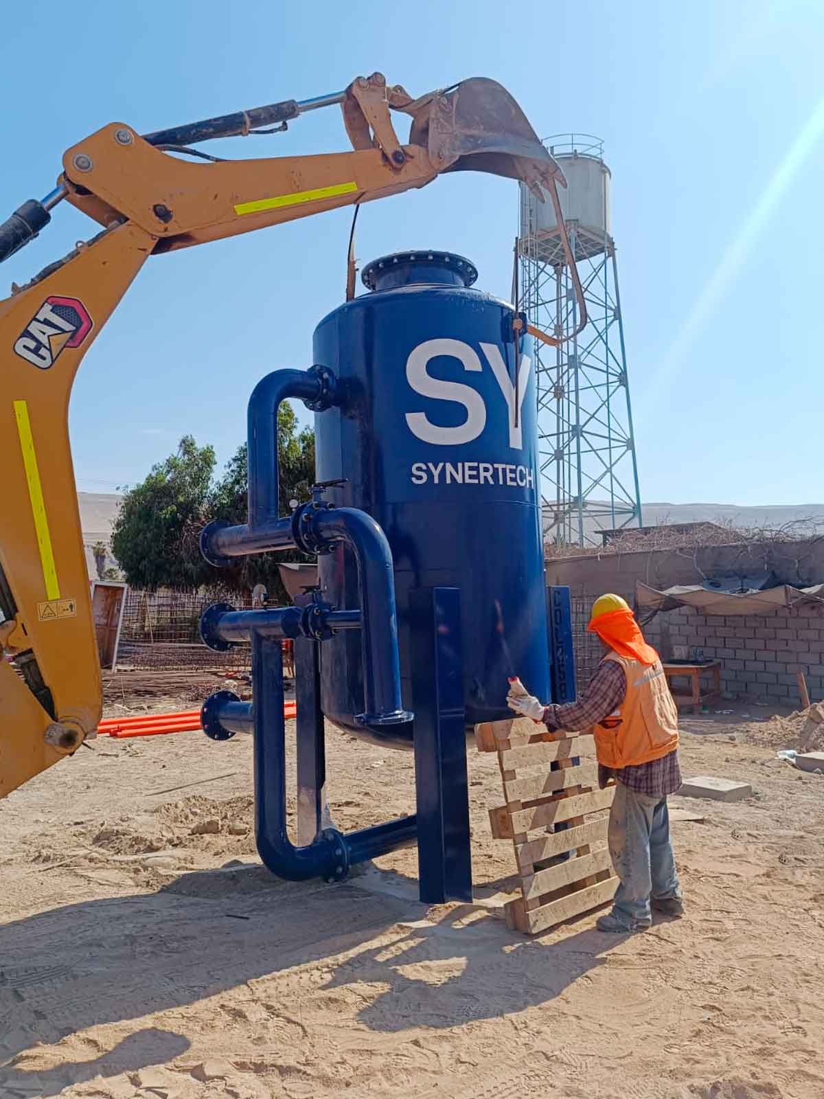 Instalación de un filtro industrial grande marcado con 'SYNERTECH' en un sitio de construcción. Una excavadora con el cubo levantado parece ayudar en el posicionamiento del filtro. Un trabajador con camiseta naranja de alta visibilidad y casco supervisa el proceso. Al fondo, se observa una estructura que parece una torre industrial, parte de la infraestructura del sitio.